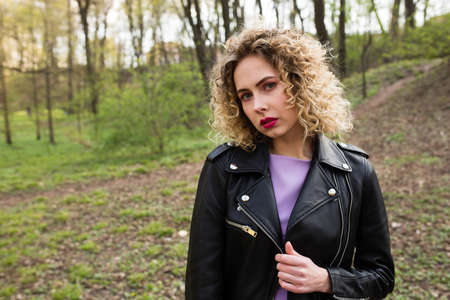 Portrait of a beautiful girl with curly long hair in black leather jacket in the spring forest