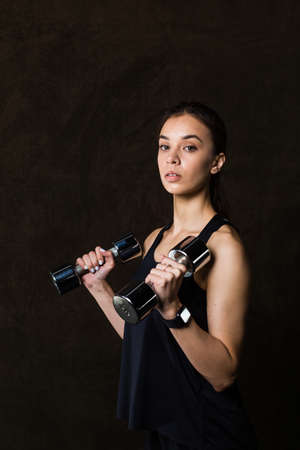 Sporty girl in fashionable sportswear holding dumbbells. Photo of muscular fitness model on dark background. Sports and Healthy lifestyleの写真素材