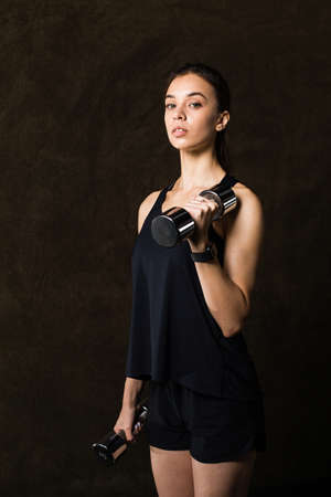 Sporty girl in fashionable sportswear holding dumbbells. Photo of muscular fitness model on dark background. Sports and Healthy lifestyleの写真素材