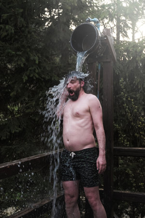 Man hardening with bucket of ice cold water after hot sauna in nature against the backdrop of the forest. concept of human emotionsの写真素材