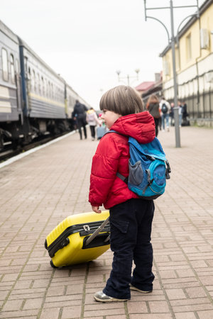small baby boy stands on the platform near the train and holds a yellow suitcase in his hands.の写真素材