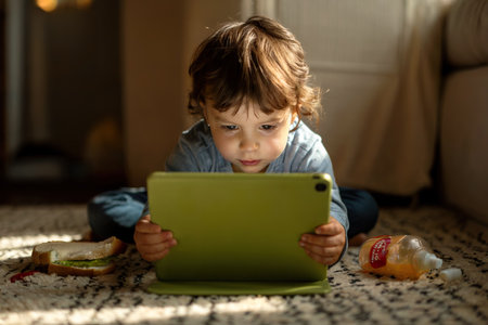 young child sitting on the floor watching videos onnline on a computer tablet while his food is left unnatended on the rug.の素材