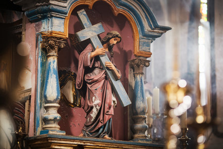 Old church setting with focus on a weathered statue of Jesus holding a cross with paint peeling and chipping. The scene is illuminated by soft window light and shows candles and church pillars in the background.の素材