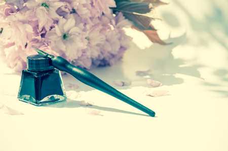 Old ink pen and ink bottle on white background. Vintage calligraphy pen and bottle of ink. Selective focus. Tender pink flowers in the background. Cherry blossom twig with black inkの写真素材