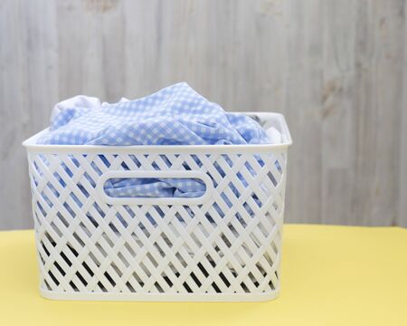 Blank blue white shirts in a white plastic basket on a yellow table on a gray background. Laundry,...