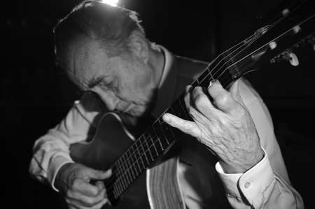 An elderly man in white shirt playing an acoustic guitar. Dark background. Monochrome.  Focus on the...