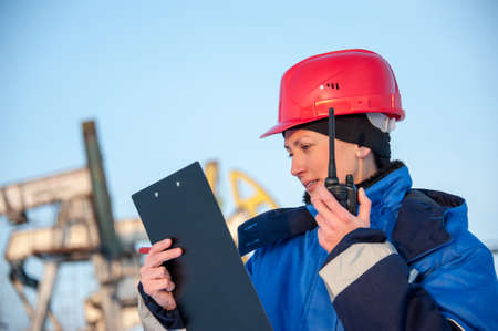 Female worker in the oil field talking on the radio wearing red helmet and blue work clothes. Industrial site background.の写真素材