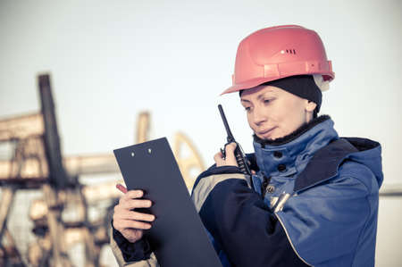 Female worker in the oil field talking on the radio wearing red helmet and blue work clothes. Industrial site background. Toned.の写真素材