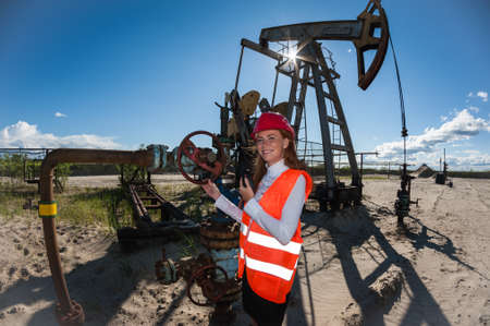 Beautiful woman engineer in the oilfield wearing red helmet and work clothes. Pump jack and wellhead valve background. Oil and gas concept.の写真素材