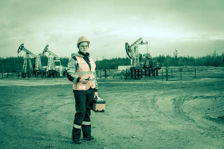Woman engineer with radio phone and toolbox in the oil field wearing orange helmet and work clothes. Industrial site background. Toned.の写真素材
