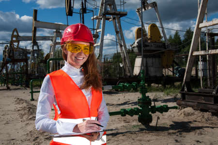 Woman engineer in yellow glasses on the oil field wearing red helmet and work clothes. Industrial site background.の写真素材