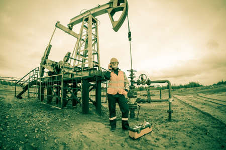 Woman engineer in the oil field talking on the radio wearing orange helmet and work clothes. Industrial site background. Toned.の写真素材