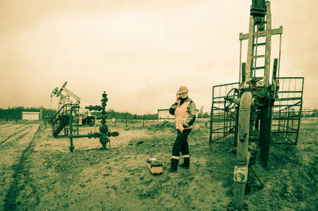 Woman engineer in the oil field talking on the radio wearing orange helmet and work clothes. Industrial site background. Toned.の写真素材