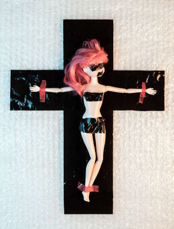 Synthetic life. Plastic doll crucified on a cross under cellophane film. Plastic pollution and social problems concept.の写真素材