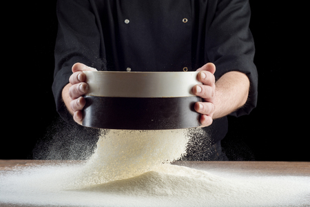 Male hands sifting flour from old sieve on old wooden kitchen table. Isolated on black background.の写真素材