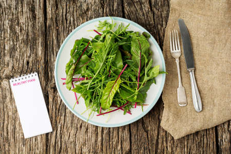 Fresh salad plate with mixed greens (arugula, mesclun, mache) on dark wooden background with cutlery and notepad. Healthy food. Green meal. Flat lay.の写真素材