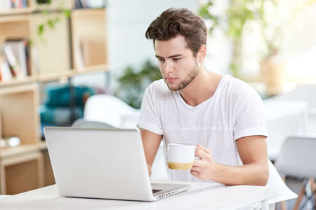 Coffee makes his day. Young handsome man drinking coffee and looking at laptop screen while sitting at his working place. Openspace, co-working, time cafeの写真素材