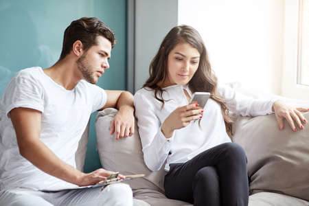 business and technology. European couple using gadgets: pretty girl in trendy clothes, typing a message on cell phone while her boyfriend sitting next to her, looking down at  her smart phone screenの写真素材