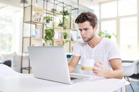 Coffee makes his day. Young handsome man drinking coffee and looking at laptop screen while sitting at his working place. Openspace, co-working, time cafeの写真素材