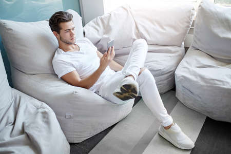 attractive young man sitting in a comfortable chair with the phone, front view. work in a modern office. Portrait of business startups. handsome guy with the phone.の写真素材