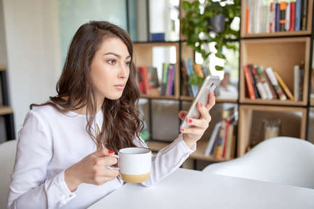 Coffee makes her day. Young handsome woman drinking coffee and looking to phone while sitting at her working place. Openspace, co-working, time cafeの写真素材