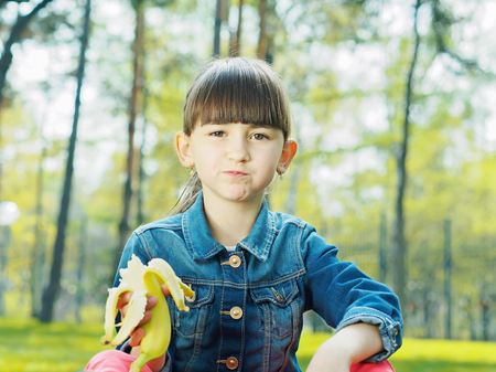 Portrait of nice caucasian girl kid eating fresh banana in summer outdoorsの写真素材