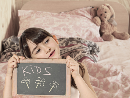 Caucasian little girl hold chalkboard signed kids with rabbit toy sitting on the bed at homeの写真素材