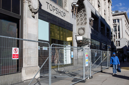 London, UK - February 26, 2021:  Barriers outside the former flagship branch of Top Shop in Oxford Circus, central London on a sunny lunchtime.  A man wearing a facemask to prevent spread of COVID 19 walks past on the virtually empty pavement.のeditorial素材