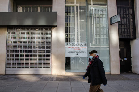 London, UK - February 26, 2021: A pedestrian wearing a face mask and protective gloves walking past a closed branch of the River Island fashion store on Oxford Street, central London.のeditorial素材