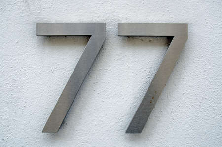 Brushed metal figures for the number seventy seven on a rough, white painted wall on the outside of...
