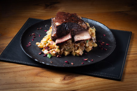 Rich, seared meat rests atop savory pasta. Red spices scatter the plate, adding contrast. Dark stone platter enhances the dish's elegance. Warm wooden table adds rustic charm
