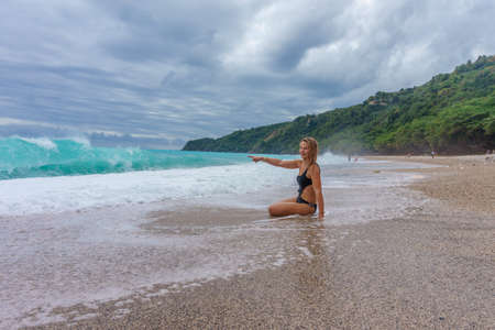 girl in bikini sits near the sea, looks at the huge waves and point to the sea with her hand. Storm in Dominican Republicの写真素材