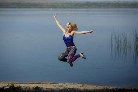 young smiling girl in colored leggings and blue top on heels jumps on a river shore. entertainment on the beach. crazy weekendの写真素材