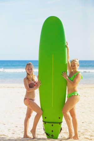 two young cute girls in bikini posing near a green surfboard on sandy beach. Welligama, Sri Lanka