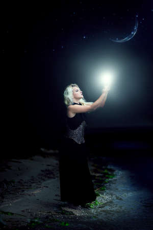 gorgeous young girl sorceress holds light at night with new moon on the skyの写真素材
