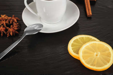 white cup of hot tea, cinnamon sticks, star anise and lemon slices on black slate square stoneの写真素材