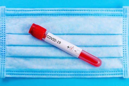 Protective medical mask with positive coronavirus blood tube test close-up on blue background....