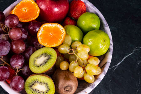 Fresh tropical fruitson in bowl on black marble background. Pineapple, papaya, mango, kiwi, grape, garnet, tangerines, bananas, apples, watermelon slices, pitaya or dragon fruitの写真素材