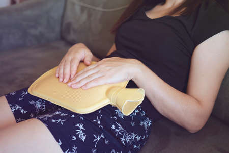 Caucasian woman lying in bed with hot water bag. Close up of woman belly with hot water bottle in bed. Beautiful young woman in bed, with hot water bag on her tummyの写真素材