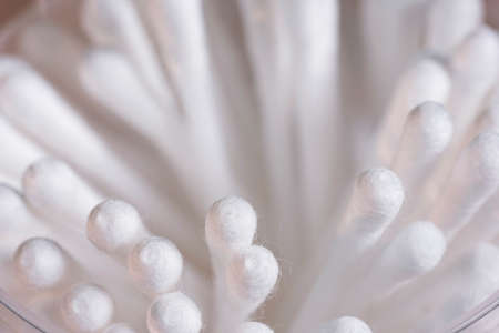white soft cotton ear sticks are stacked in a small pile on a white surface. the container is...