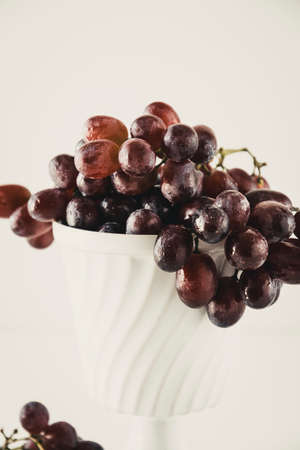 Dark red grapes in an old white porcelain bowl standing on a white table with some grapes lying on the table in the foreground. Close-up.の写真素材