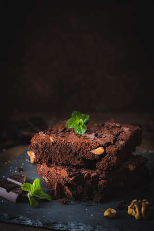Stacked pieces of walnut brownies on a slate platter and dark brown background, decorated with walnuts, chocolate and mint leaves, vertical with copy spaceの写真素材