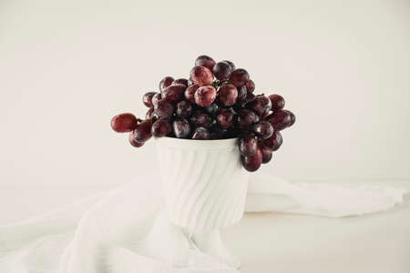 Dark red grapes in an old white porcelain bowl standing on a white table with a white cloth draped around.の写真素材