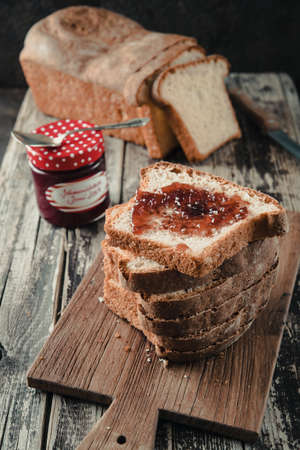 Stacked slices of homemade white bread lying on a wooden board, the uppest spread with jam, the chunk of bread and the glass with jam in the backgroundの写真素材