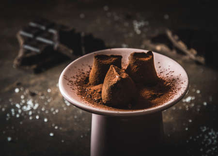 Three chocolate pralines with powdered cocoa lying on a pink plate, with dark brown chocolate in dark background, closeupの写真素材