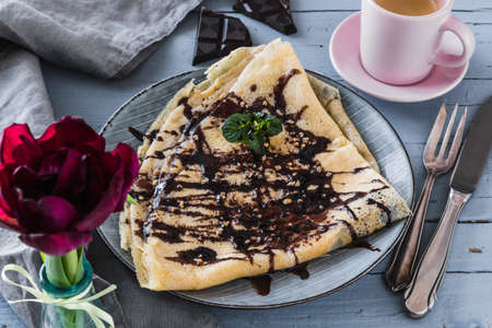 French pancake with chocolate, brittle and mint, decorated with espresso in a rose cup, dark red tulip, gray napkin and cutlery lying on a light gray wooden table.の写真素材