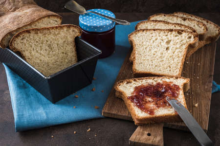 Slices of homemade white bread lying on a wooden board and blue napkin, the front spread with jam, the chunk of bread and the glass with jam in the background. Vertical with copy space.の写真素材