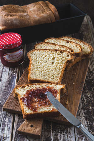 Slices of homemade white bread lying on a wooden board on a wooden table, the front spread with jam,...