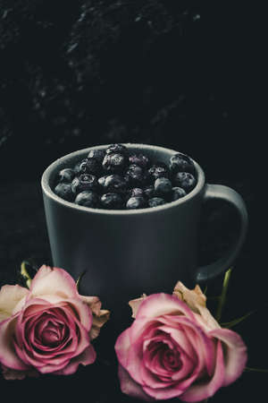 Blue-gray cup filled with ripe blueberries, decorated with two roses on dark background with copy space.の写真素材