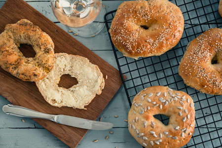 Homemade bagels on a cooling rack on gray wood, one with butter on a wooden board, glass of wine. Top view.の写真素材